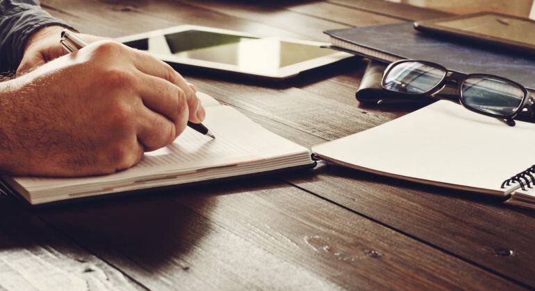 Imagen de un hombre escribiendo junto a un dispositivo digital, varias libretas y un par de gafas.