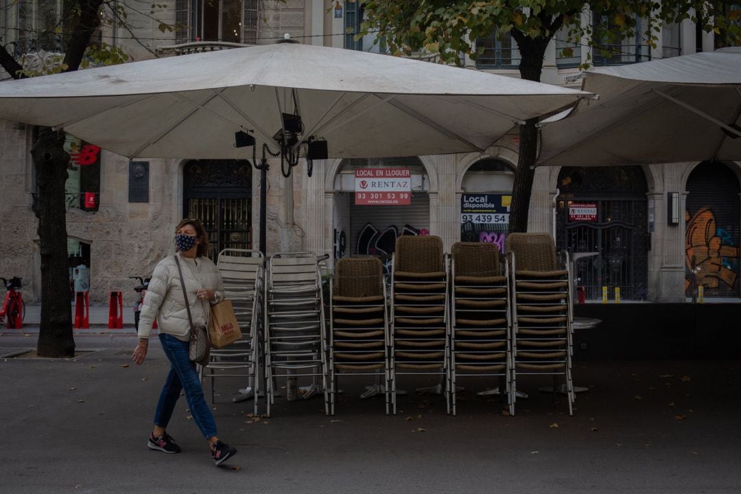 Una mujer pasa junto a la terraza recogida de un bar cerrado.