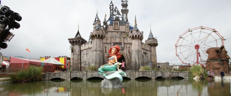 El palacio de Dismaland parodia al de Disney, al igual que lo hace la sirenita distorsionada sobre un estanque enfanganado. 