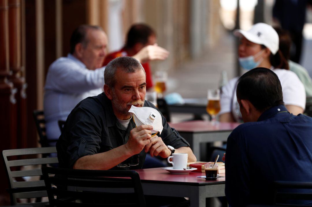 Varios ciudadanos tomando café, uno de ellos enseña su mascarilla de protección 