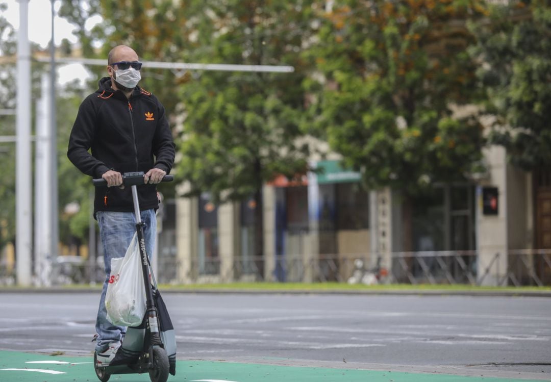 Un joven con mascarilla transporta su compra en Sevilla usando un patinete 