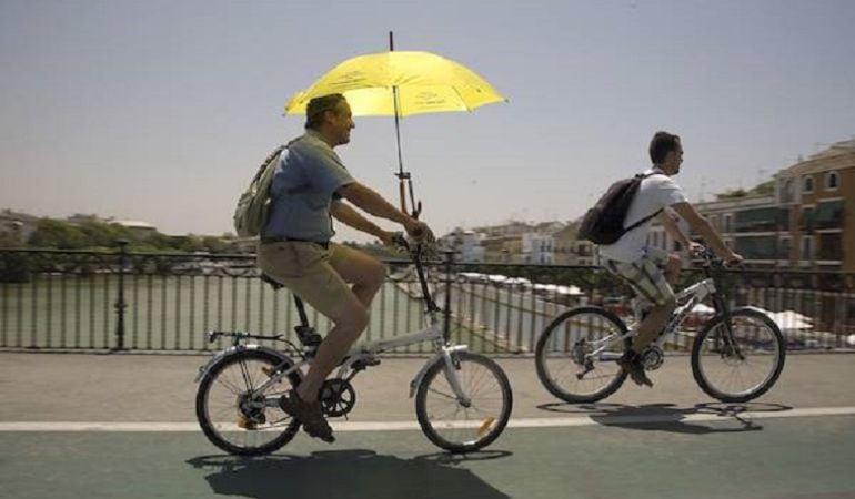 Un ciclista cruza el Puente de Triana cobijado bajo una sombrilla