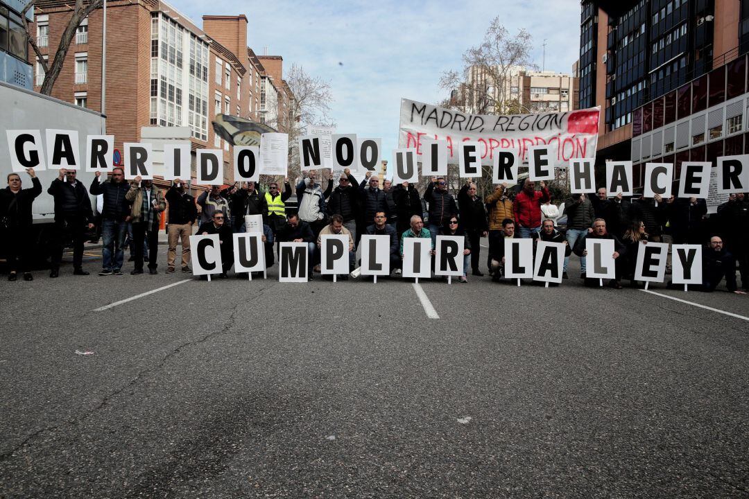 Algunos de los taxistas concentrados frente a la Consejería de Transportes, Movilidad e Infraestructuras para exigir que se &quot;cumpla la ley&quot; respecto a los vehículos de transporte con conductor (VTC), sujetan carteles contra el consejero Ángel Garrido.